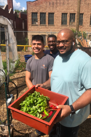 Picture of three men outdoors on a sunny day, smiling at the camera. the man in the foreground is holding a red box with plant seedlings in it. A red bridk building can be seen in the background. A garden hoop house is seen at left. 