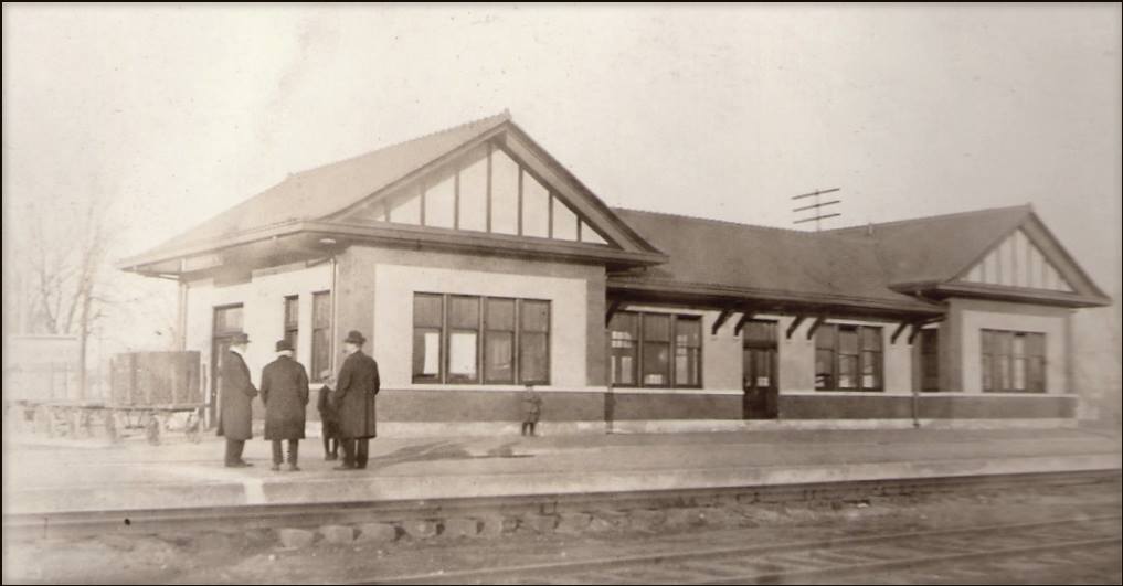 Black and white picture of the outside of a train depot, with passengers waiting on the platform