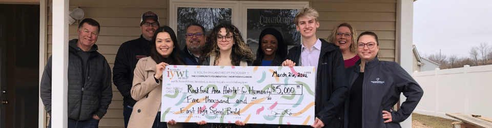 Group of smiling people standing on the front porch of a house under construction, holding a large check. 